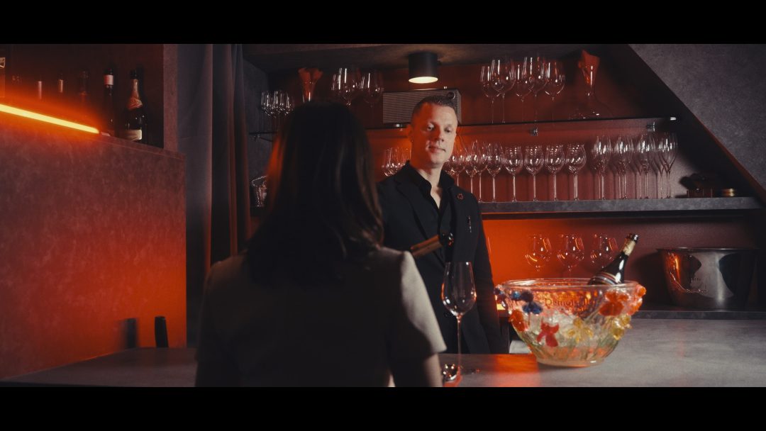 Bartender and guest in conversation at bar, shelves of glasses behind.