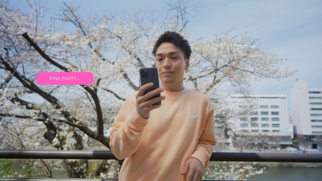 Man in peach sweater holding phone with cherry blossoms behind.