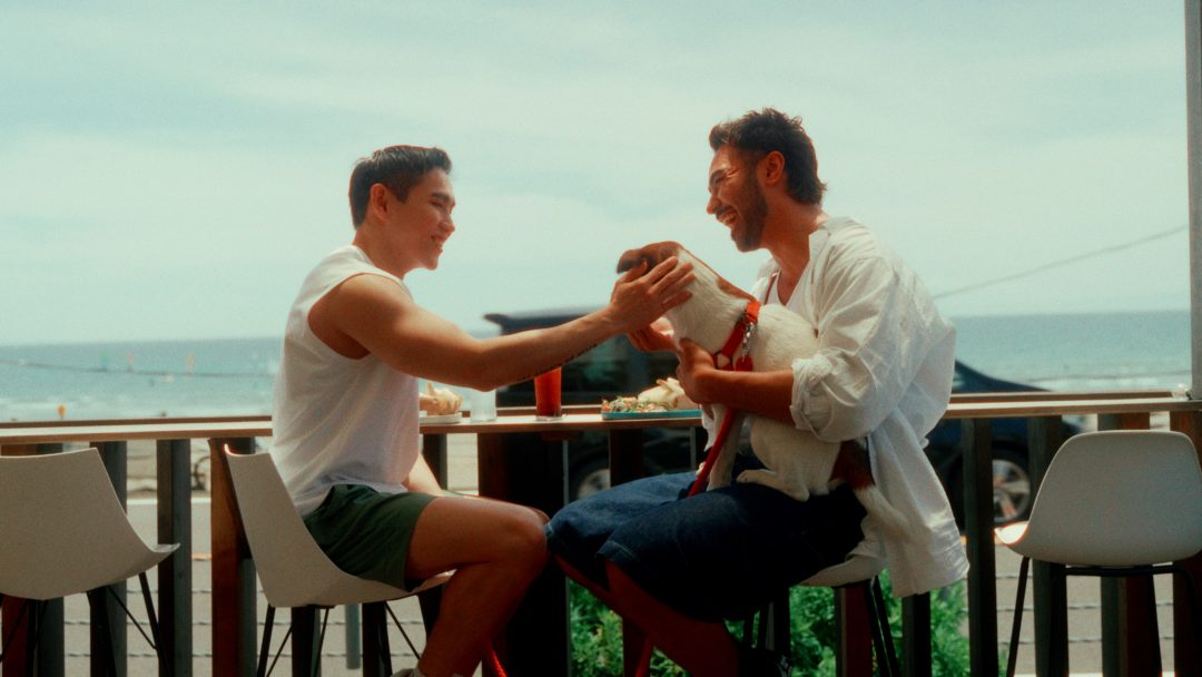 Two men laughing and sharing food at outdoor seaside café.