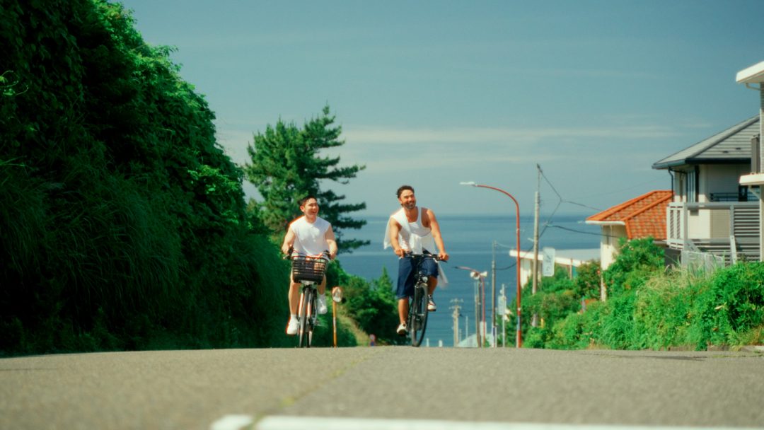Two men cycling uphill near seaside houses on sunny day.