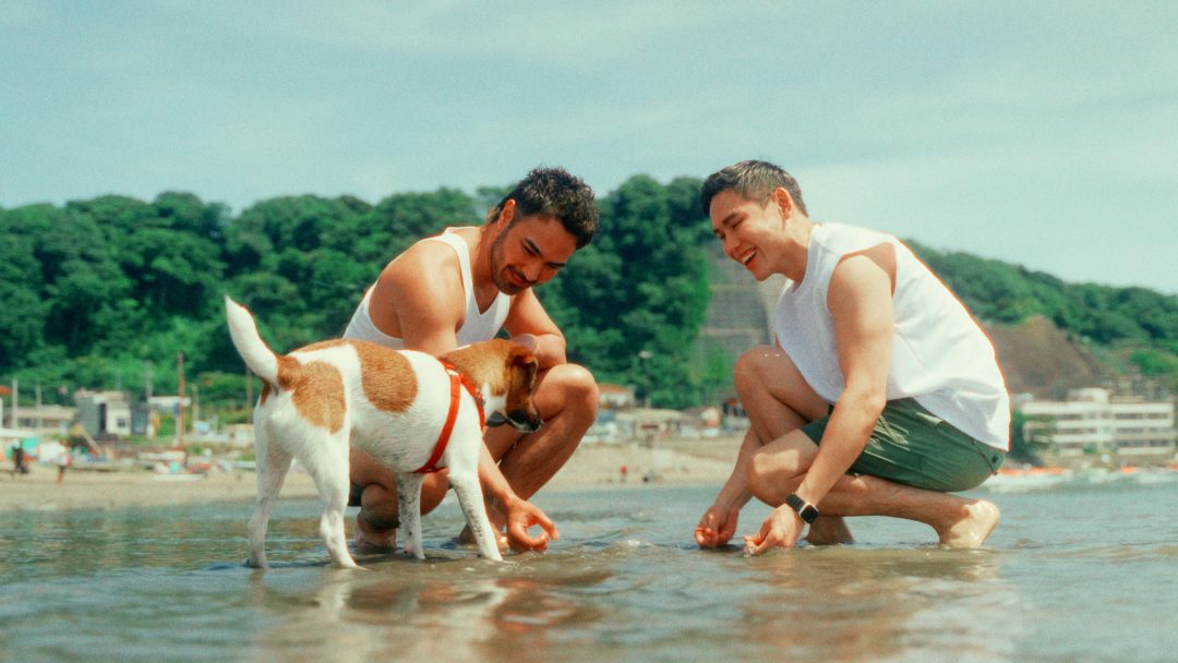 Two men crouching by the sea petting a dog in shallow water.