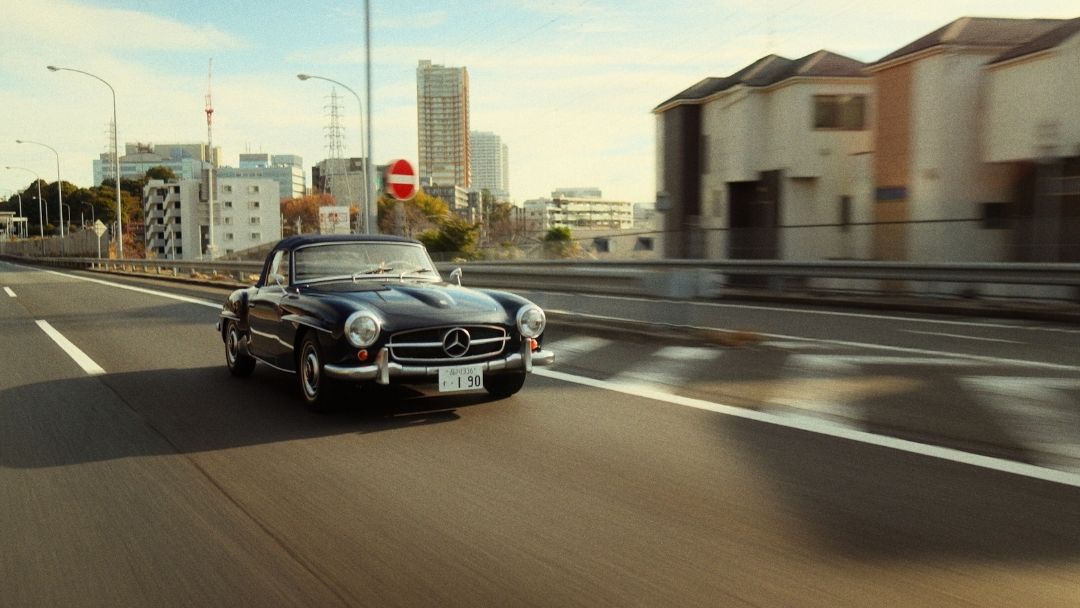 Mercedes 190SL driving fast on Tokyo expressway skyline behind.